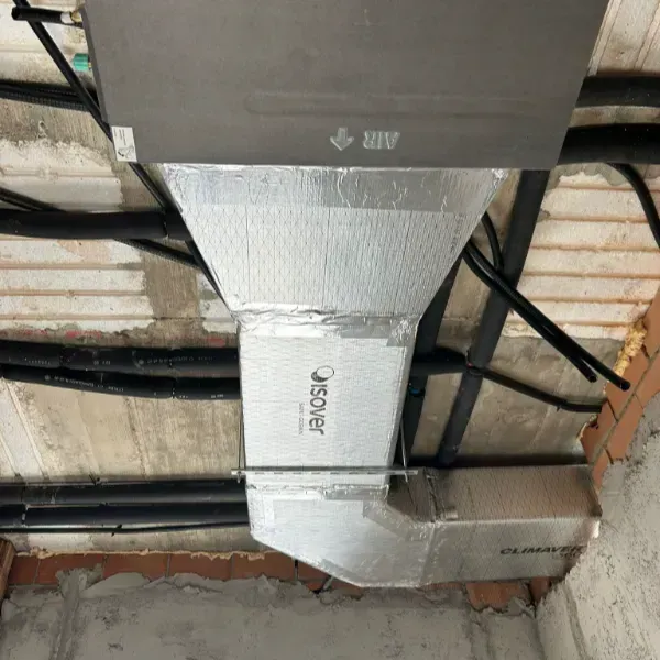 Conductos de HVAC aislados, envueltos con papel de aluminio plateado, instalados en un techo, tuberías negras visibles.