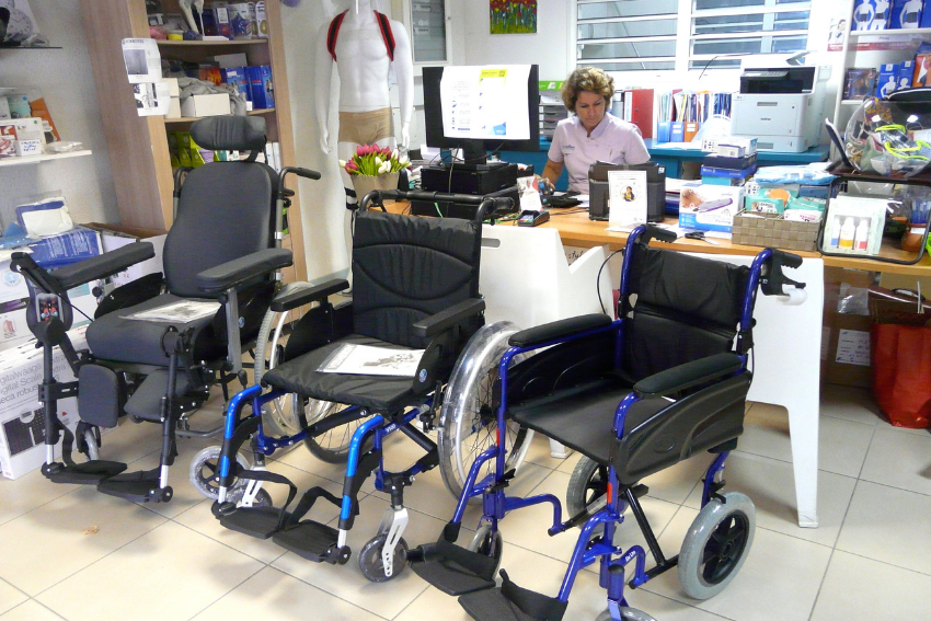 Trois fauteuils roulants sont exposés dans un magasin ; une personne derrière un comptoir est visible en arrière-plan.