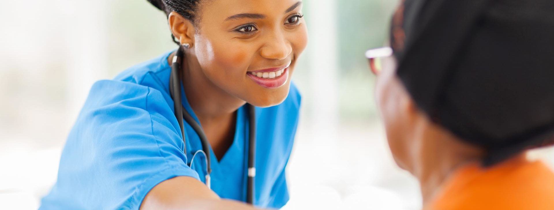 Une soignante souriante en blouse bleue se penche vers un patient vêtu d'un haut orange.