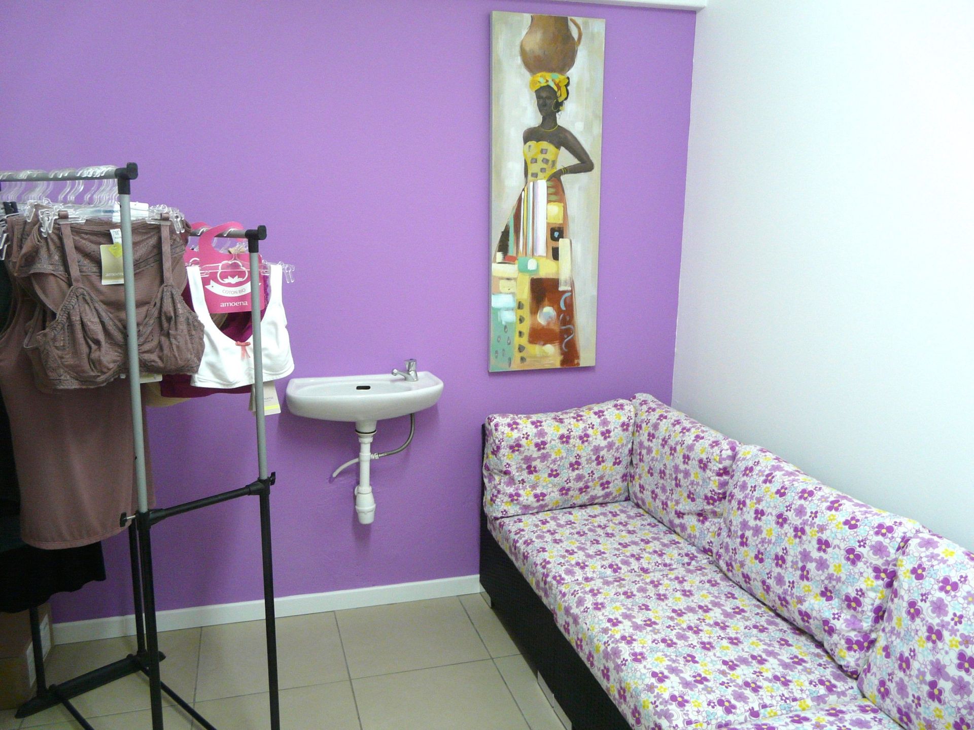 Petite pièce avec un mur violet, un canapé, un lavabo, un portant à vêtements et un tableau représentant une femme.