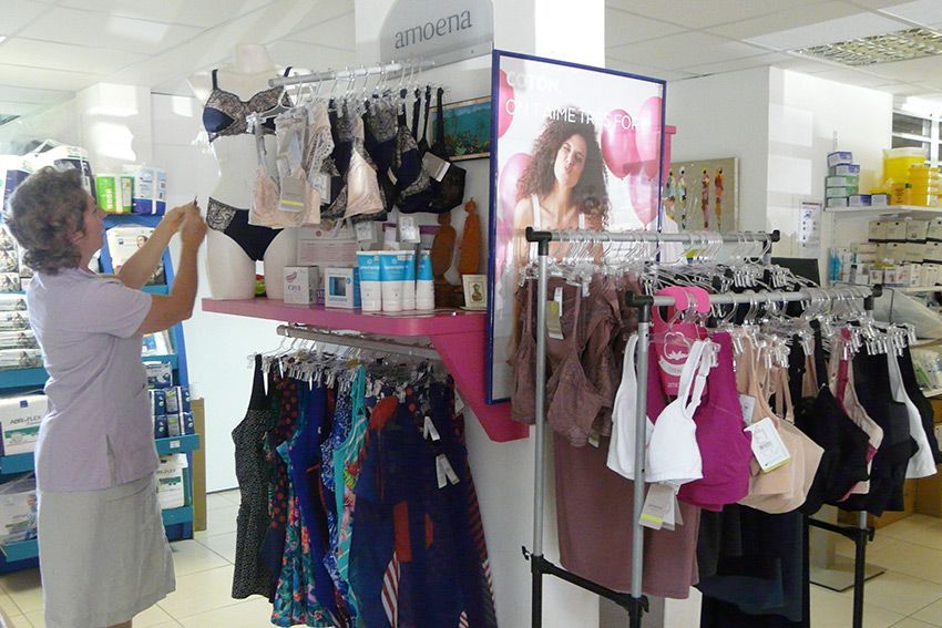 Une femme dispose de la lingerie sur un présentoir de magasin. Différents soutiens-gorge et sous-vêtements sont suspendus à des portants. Éclairage vif, ambiance de magasin.