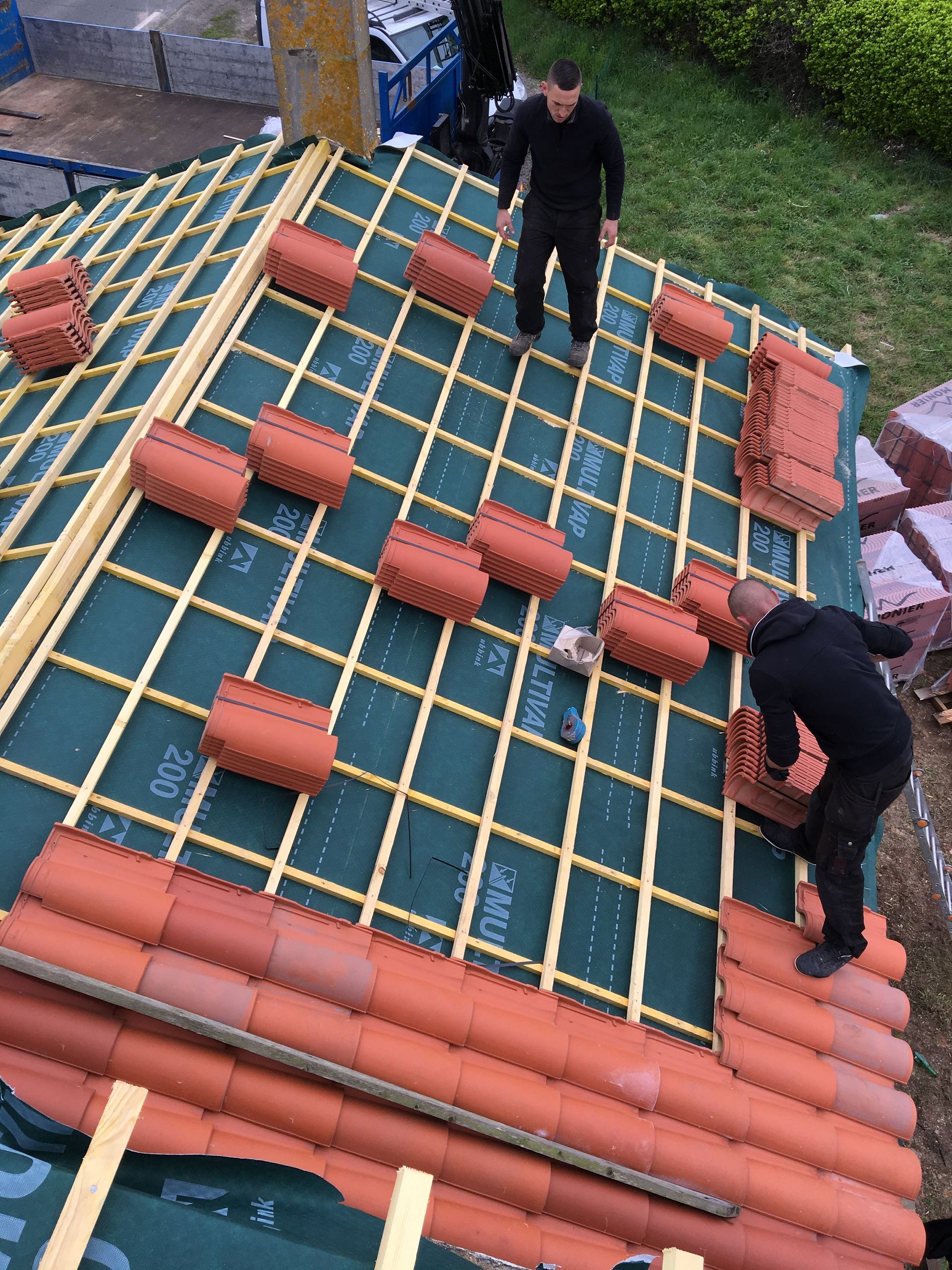 Des couvreurs posent des tuiles en terre cuite sur un toit, avec des supports en bois et une sous-couche verte.