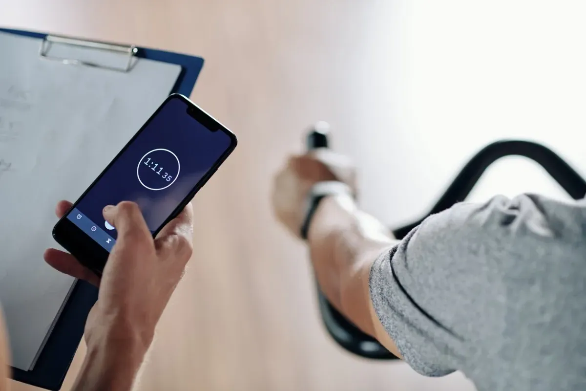 Persona que usa el teléfono para cronometrar su entrenamiento en bicicleta estática; portapapeles visible.