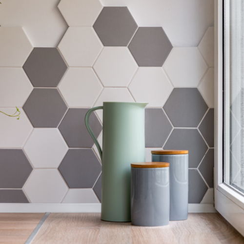 Trois bidons et un pichet sur une surface en bois, avec un mur de carreaux hexagonaux dans des tons de gris et de blanc.