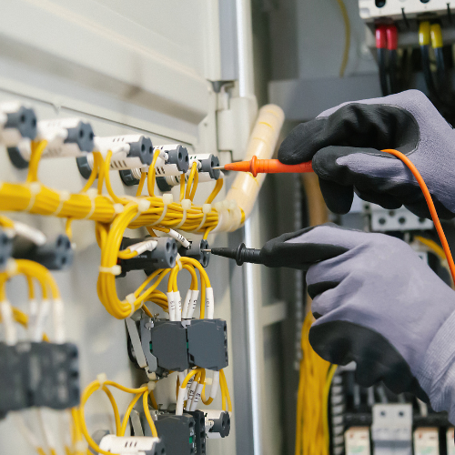 Des mains gantées testent des fils électriques dans une armoire avec un multimètre. Fils jaunes et noirs visibles.