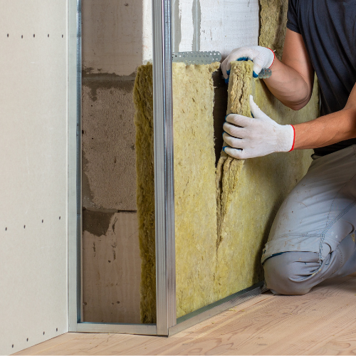 Une personne installe de l'isolant entre des montants métalliques. Elle porte des gants et l'isolant est jaune-brun. =