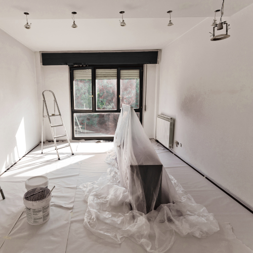 Une pièce en rénovation. Murs blancs, meubles recouverts, échelle, pots de peinture et fenêtre donnant sur l'extérieur.
