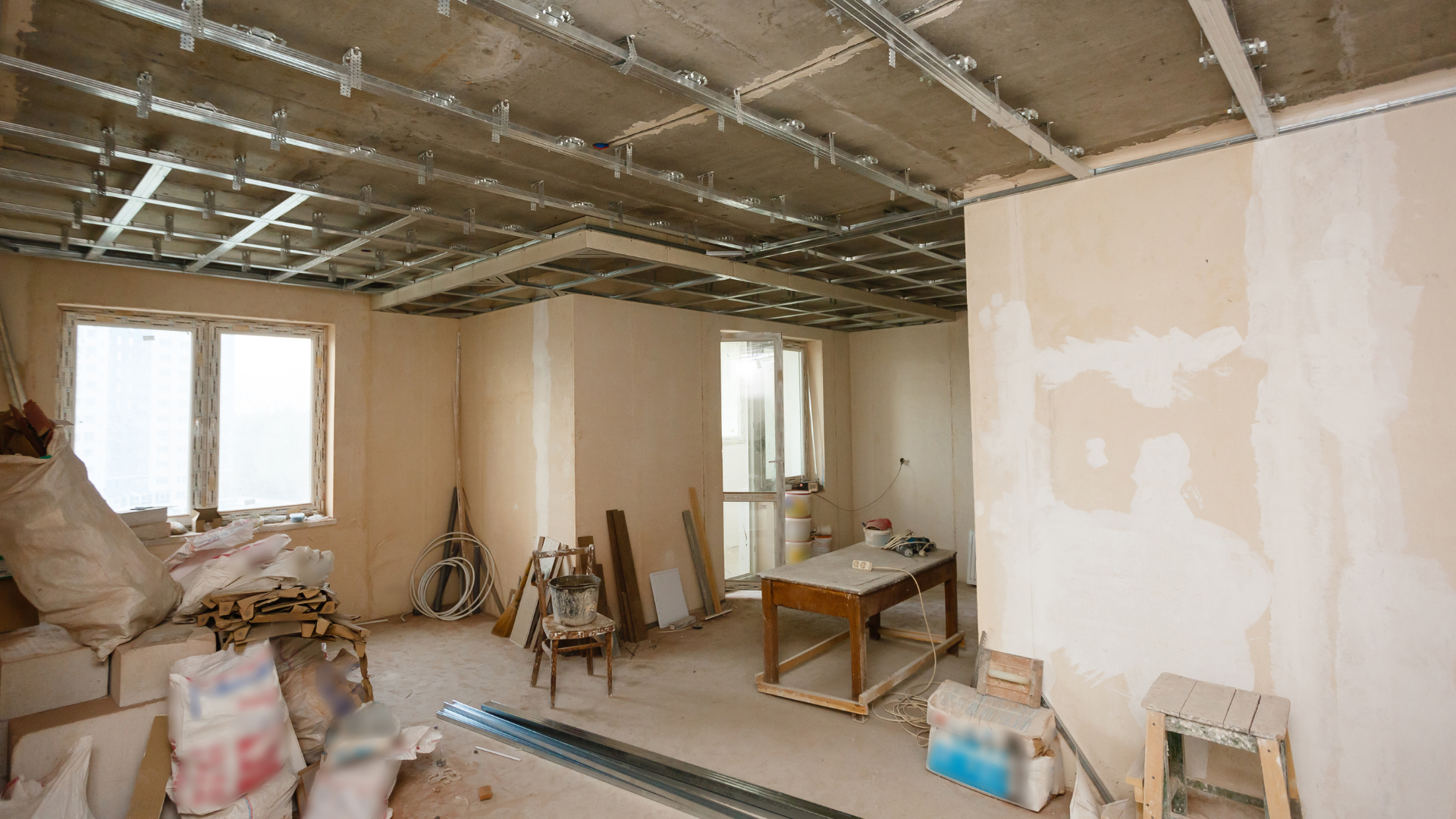 Pièce intérieure en construction avec murs non finis, grille de plafond apparente, fenêtres et matériaux de construction.