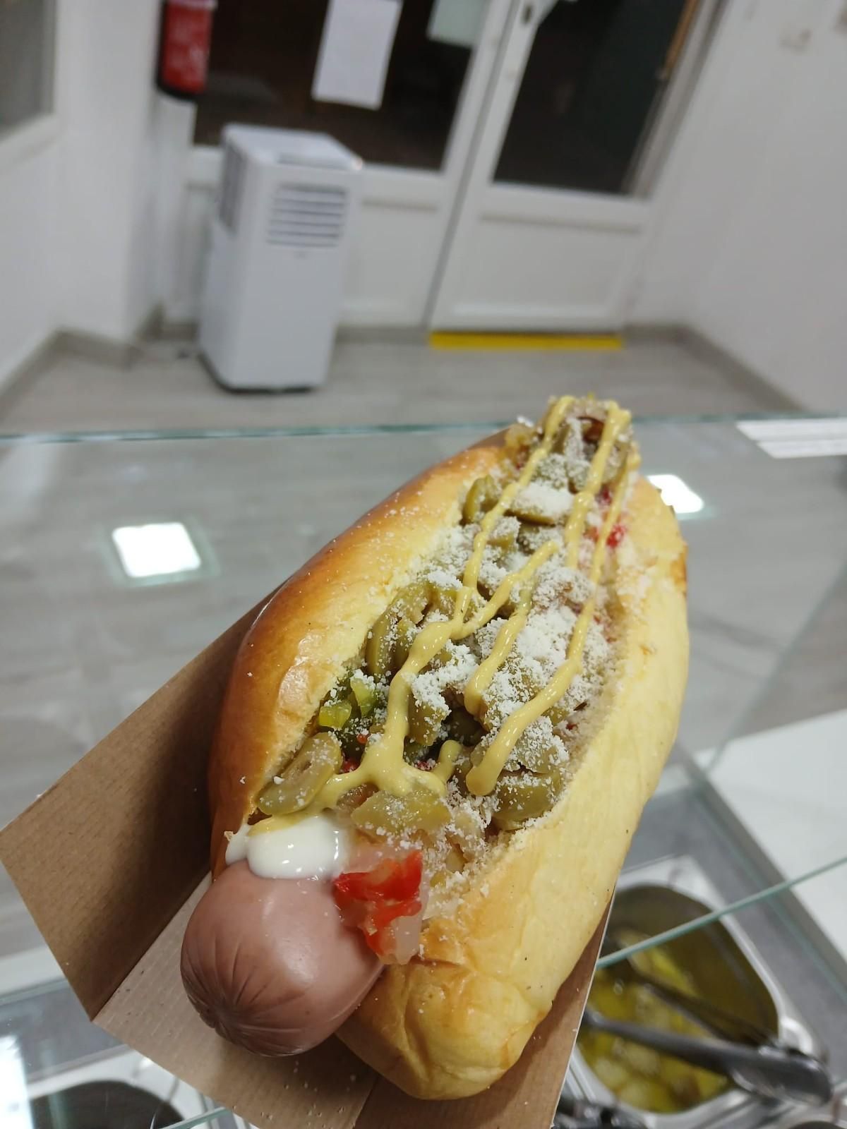 Perro caliente en un pan, cubierto con salsa verde, mostaza y queso, sobre un papel marrón, dentro de una tienda.
