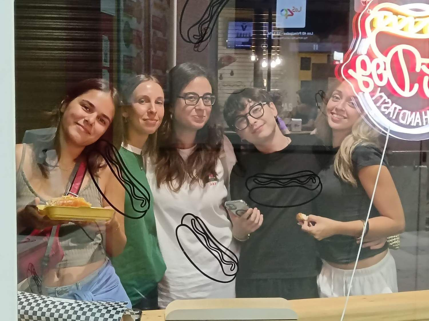 Cinco personas sonríen tras una ventana. Una sostiene una bandeja de comida