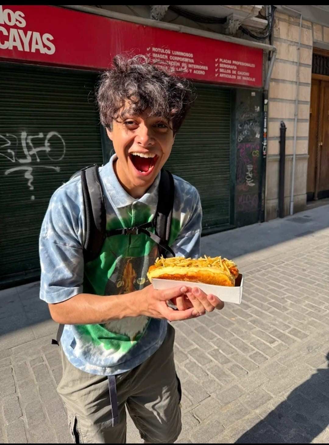 Persona con un perrito caliente en la calle, sonriendo ampliamente. Lleva una camiseta tie-dye y una mochila.