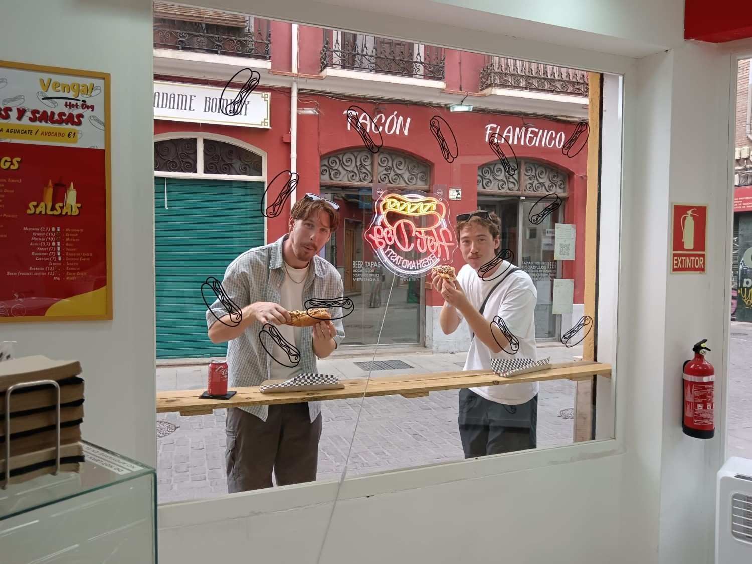 Dos personas comiendo en un mostrador junto a una ventana, con una calle y negocios visibles afuera.