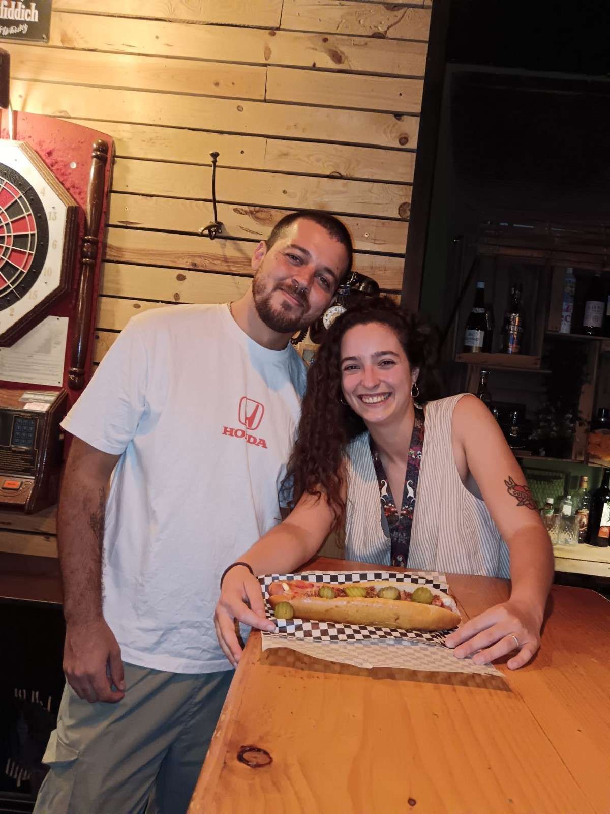 Hombre y mujer en un bar, con un sándwich en la mano. El hombre sonríe, la mujer lo mira.