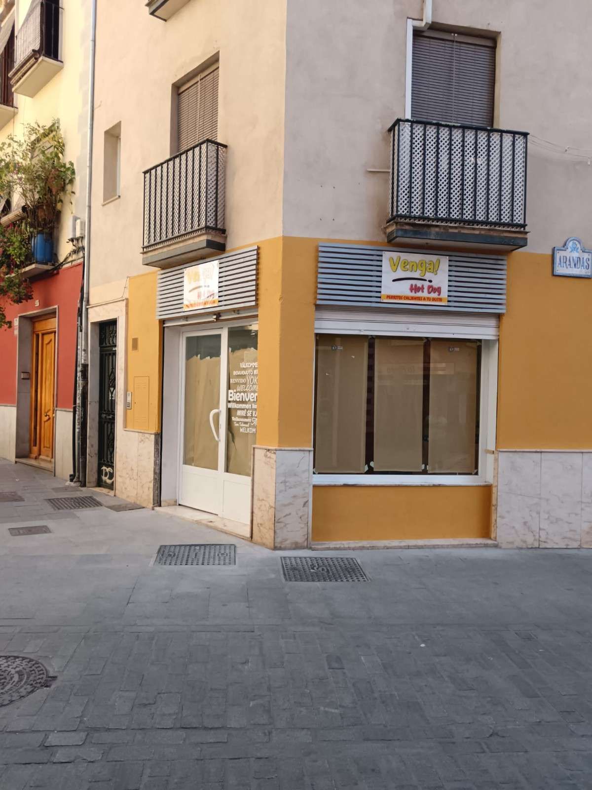 Local comercial en esquina con fachada en tonos tostados y amarillos, con pequeños balcones en la parte superior. 