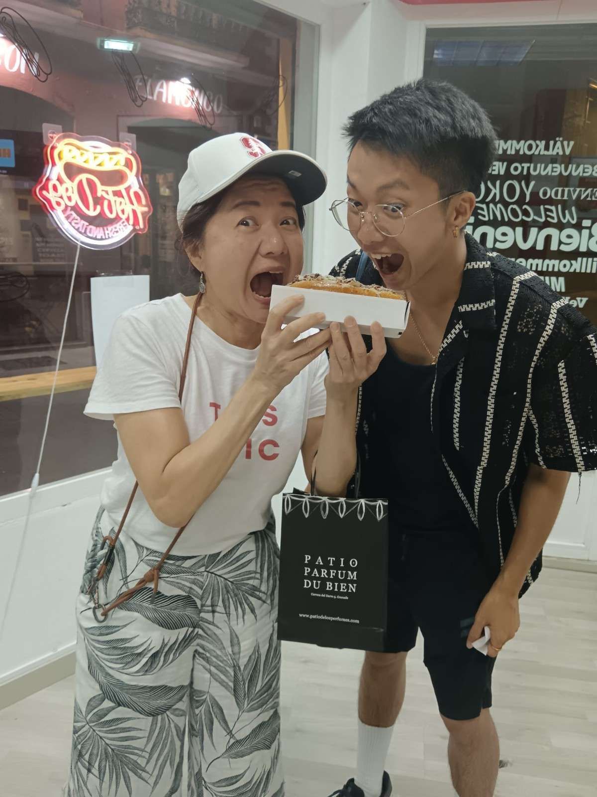 Dos personas comen de una caja de comida para llevar. Tienen cara de emoción y están dentro de una tienda bien iluminada.