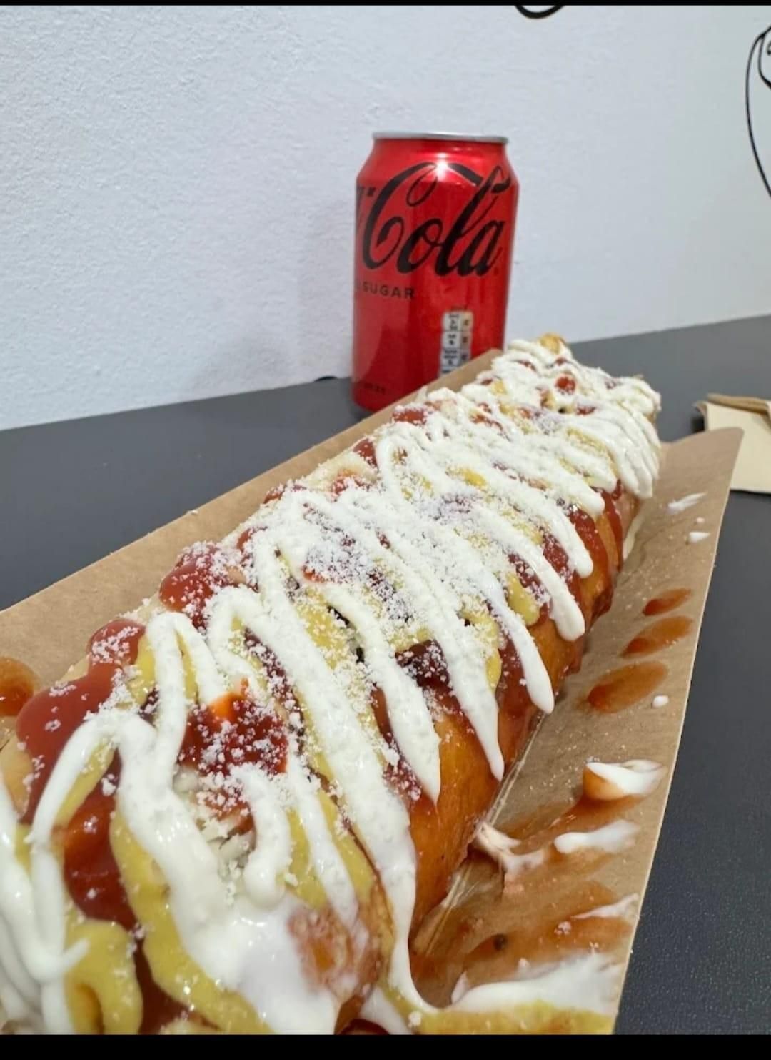 Un gran hot dog con aderezos y una lata de Coca-Cola sobre una mesa.