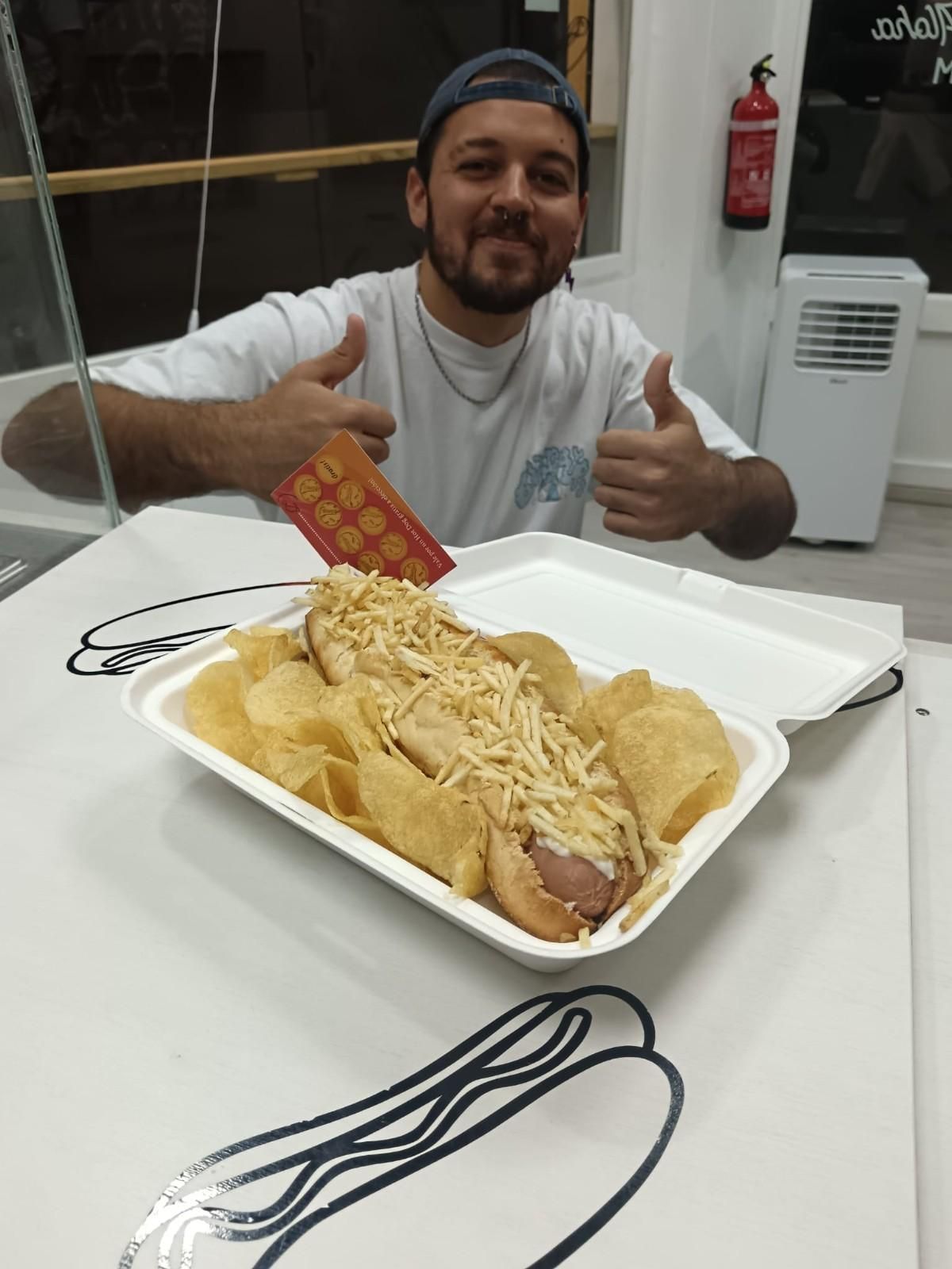 Hombre sonriendo, haciendo un gesto de aprobación con el pulgar, junto a un envase de hot dog