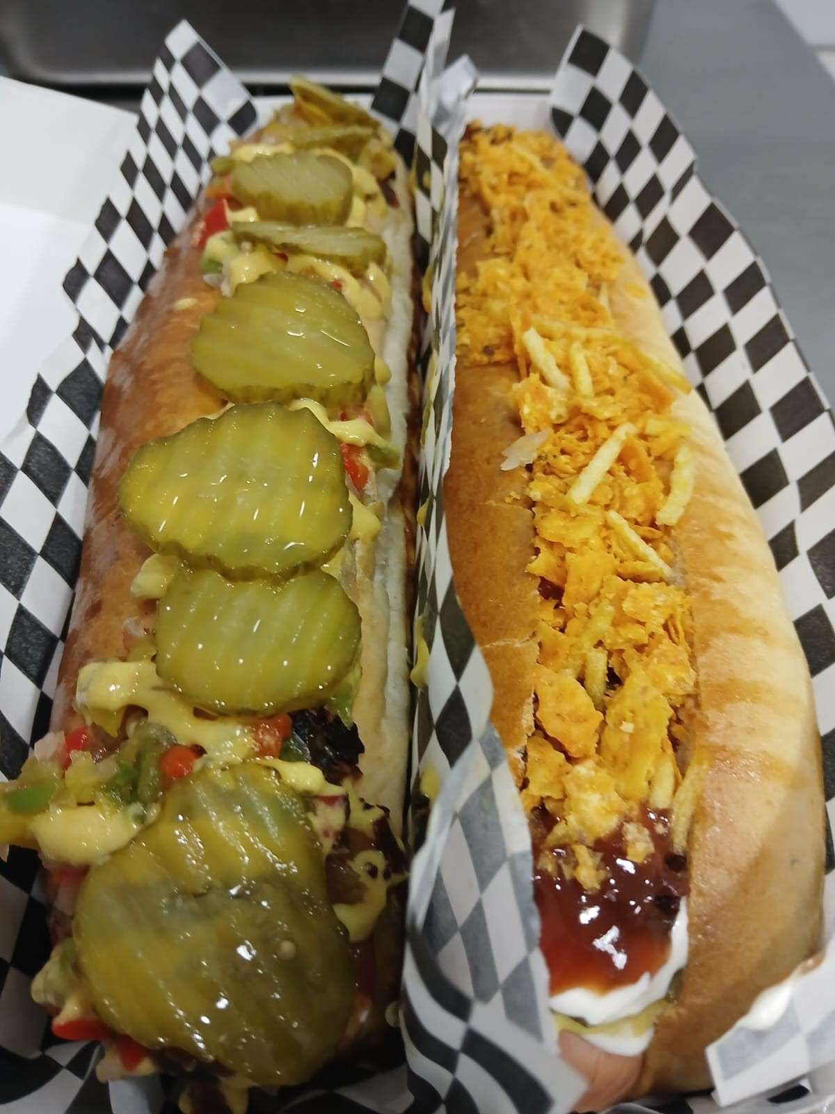 Dos perritos calientes completamente cargados en recipientes de papel a cuadros: uno con pepinillos 