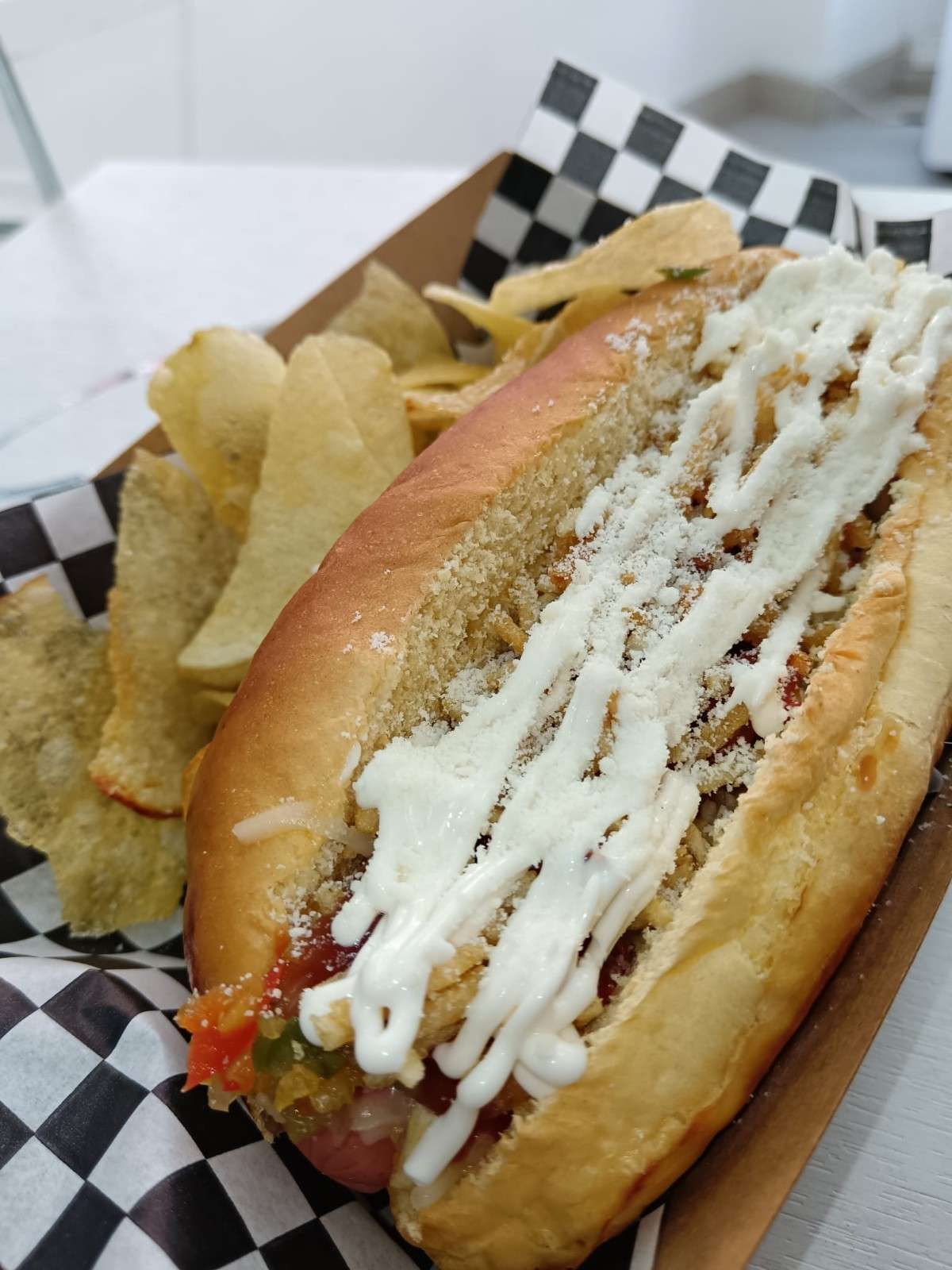 Perro caliente con aderezos y patatas fritas en una cesta, sobre un revestimiento a cuadros.