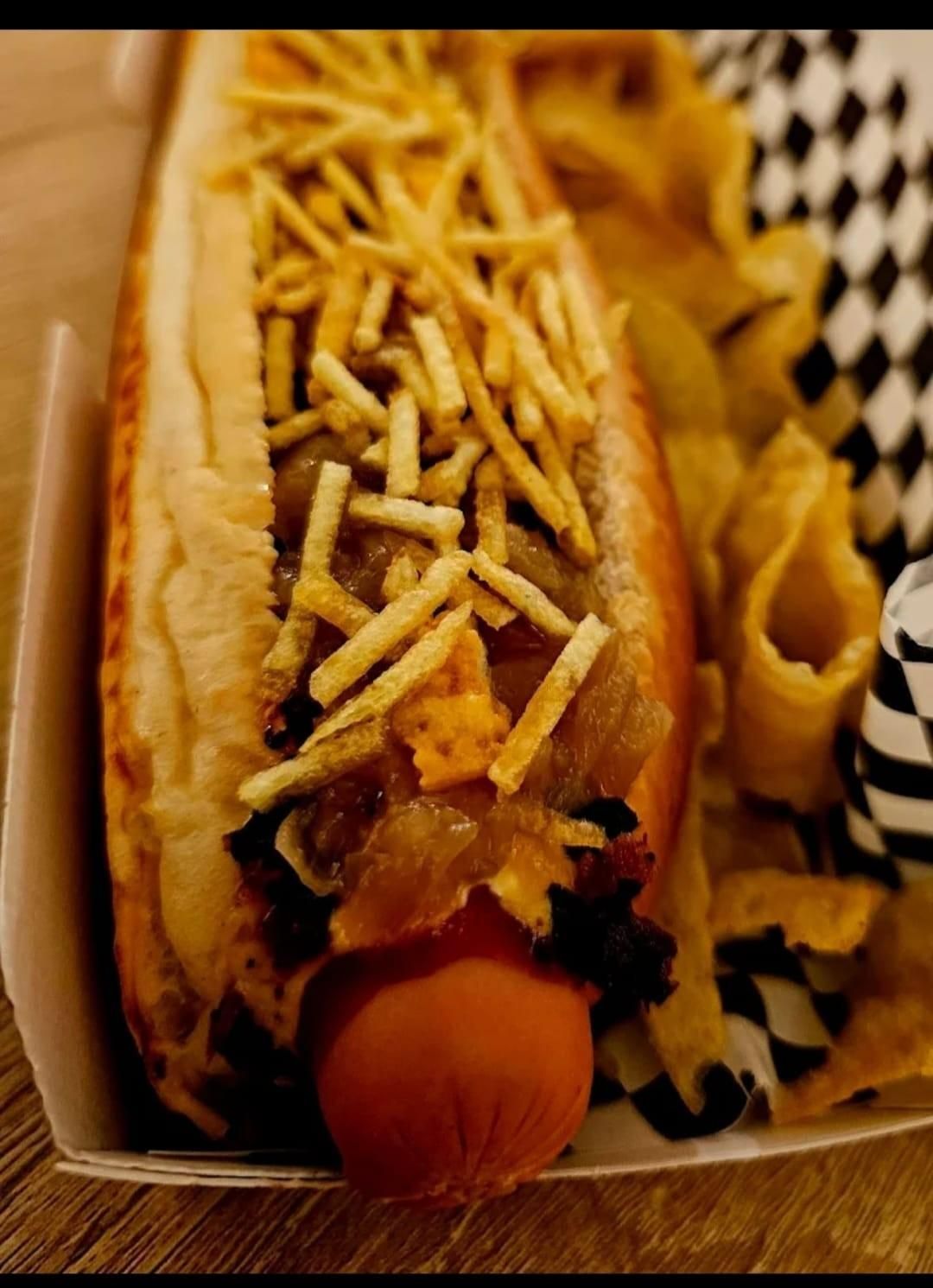 Perro caliente en pan, cubierto con papas fritas crujientes y cebollas, servido con papas fritas en un recipiente de papel.