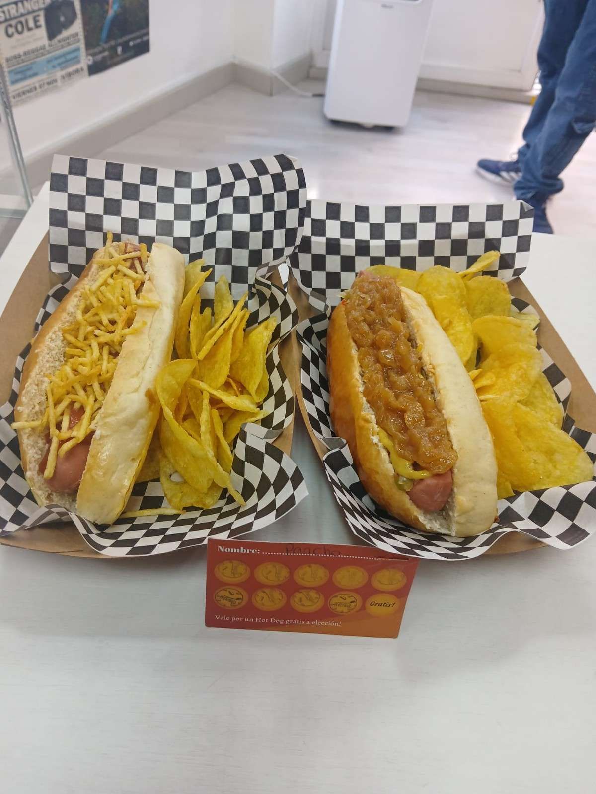 Dos perritos calientes con aderezo y patatas fritas en bandejas de papel a cuadros.