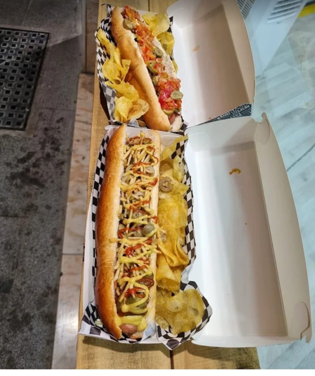 Dos hot dogs en cajas abiertas con papas fritas. Uno con varios ingredientes, el otro con salsa.