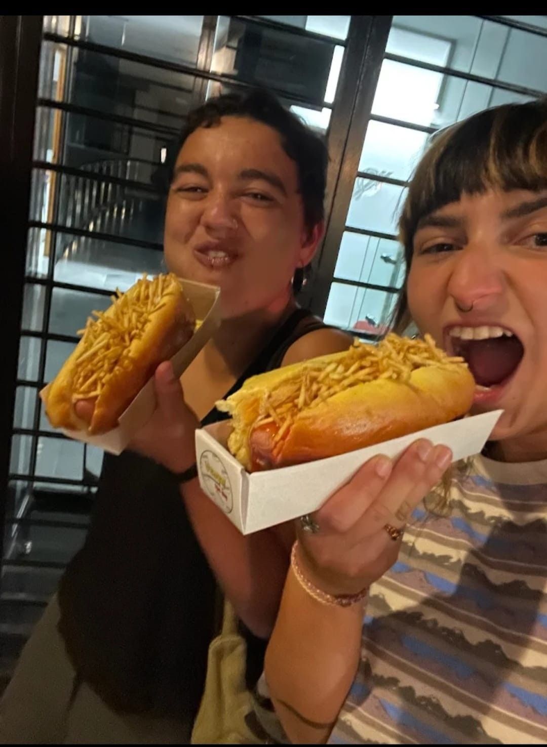 Dos personas con perritos calientes y papas fritas, una a medio morder. Al aire libre, sonriendo.