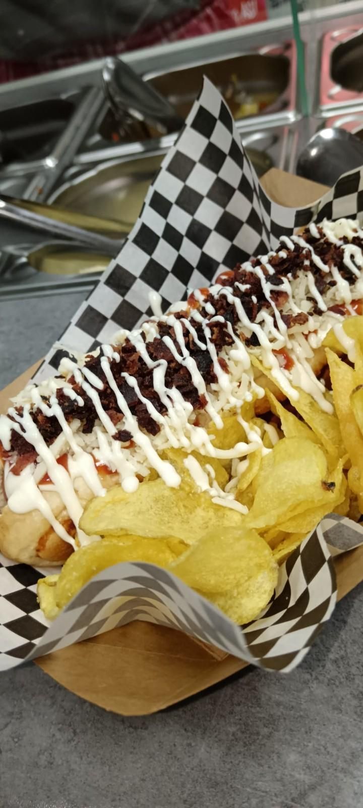 Perro caliente envuelto en papel a cuadros, cubierto con tocino, salsa blanca y servido con patatas fritas.