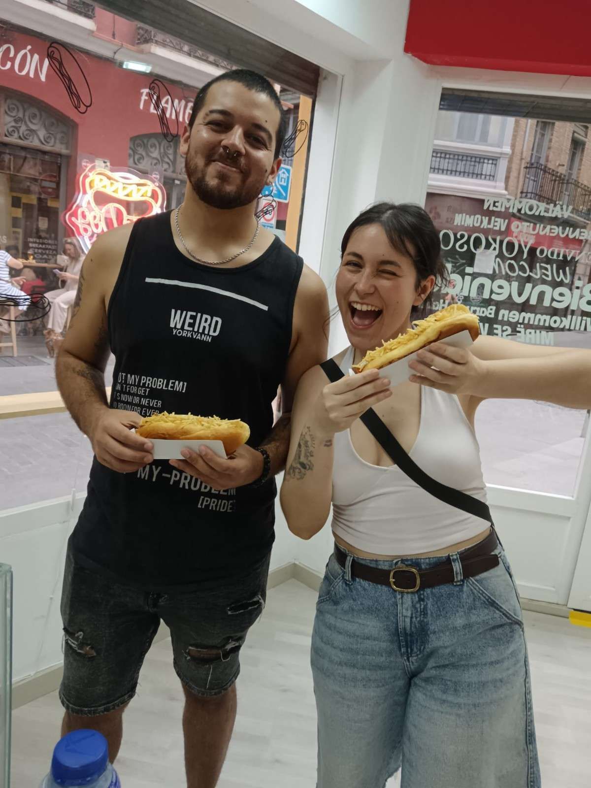 Dos personas sosteniendo sándwiches grandes, sonriendo en una tienda.