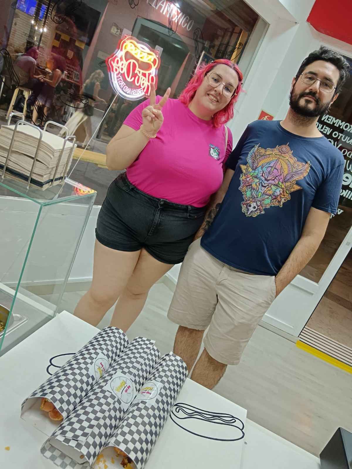 Una mujer con camisa rosa hace la señal de paz, un hombre a su lado, con envoltorios de comida sobre la mesa. 