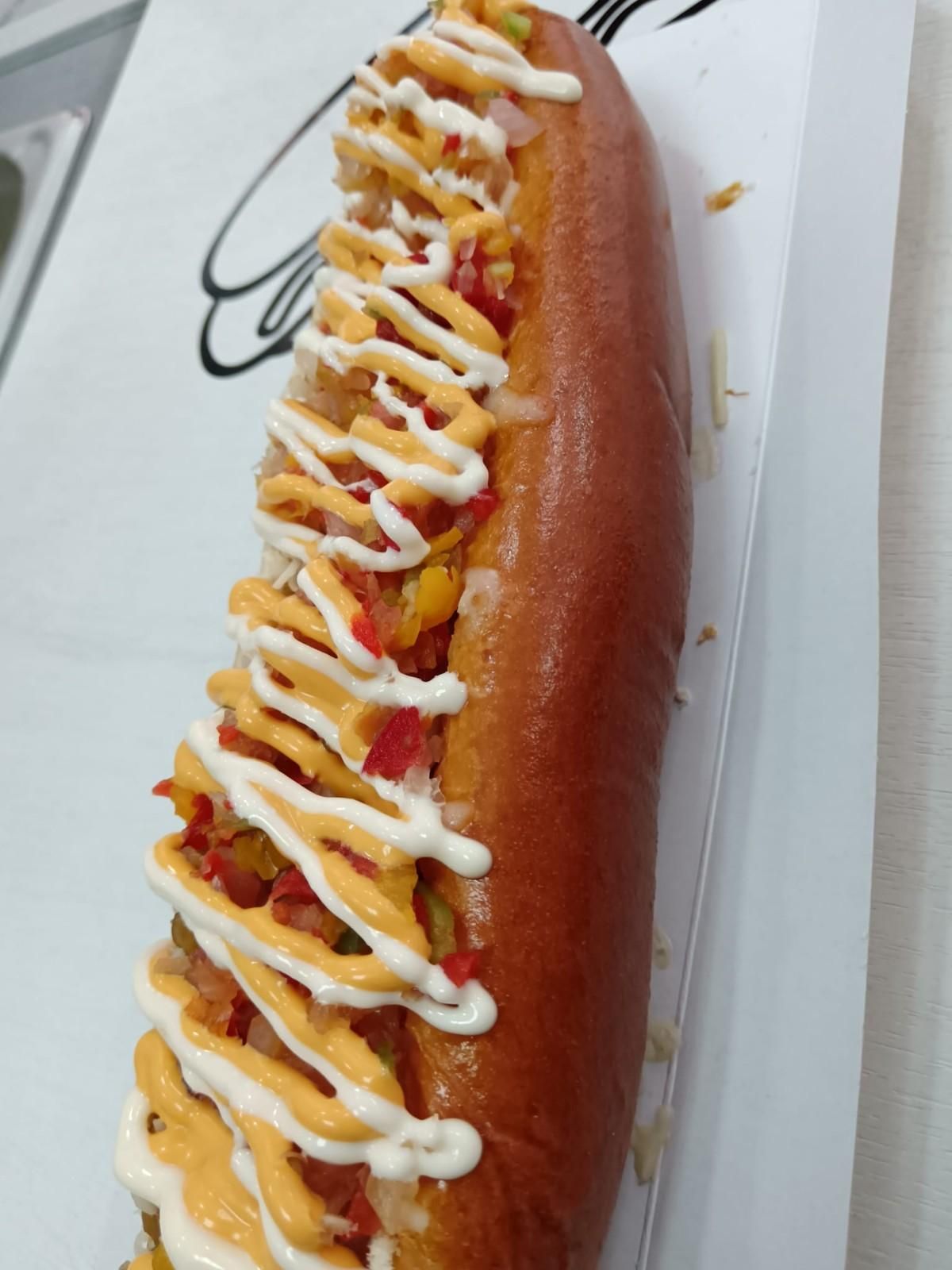 Perro caliente en pan, cubierto con verduras cortadas en cubitos y rociado con queso y salsa blanca.