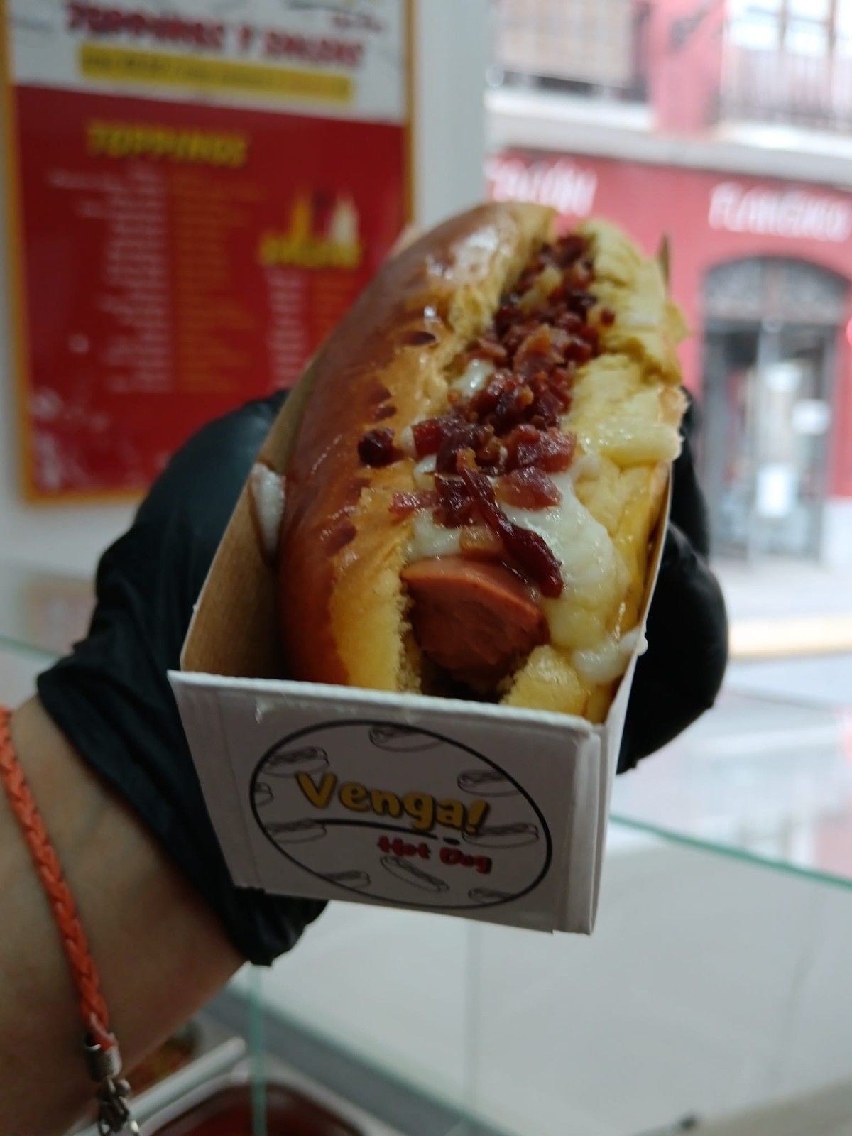 Un perrito caliente en una caja, sostenido por una mano enguantada. Incluye tocino y queso. Un cartel al fondo.