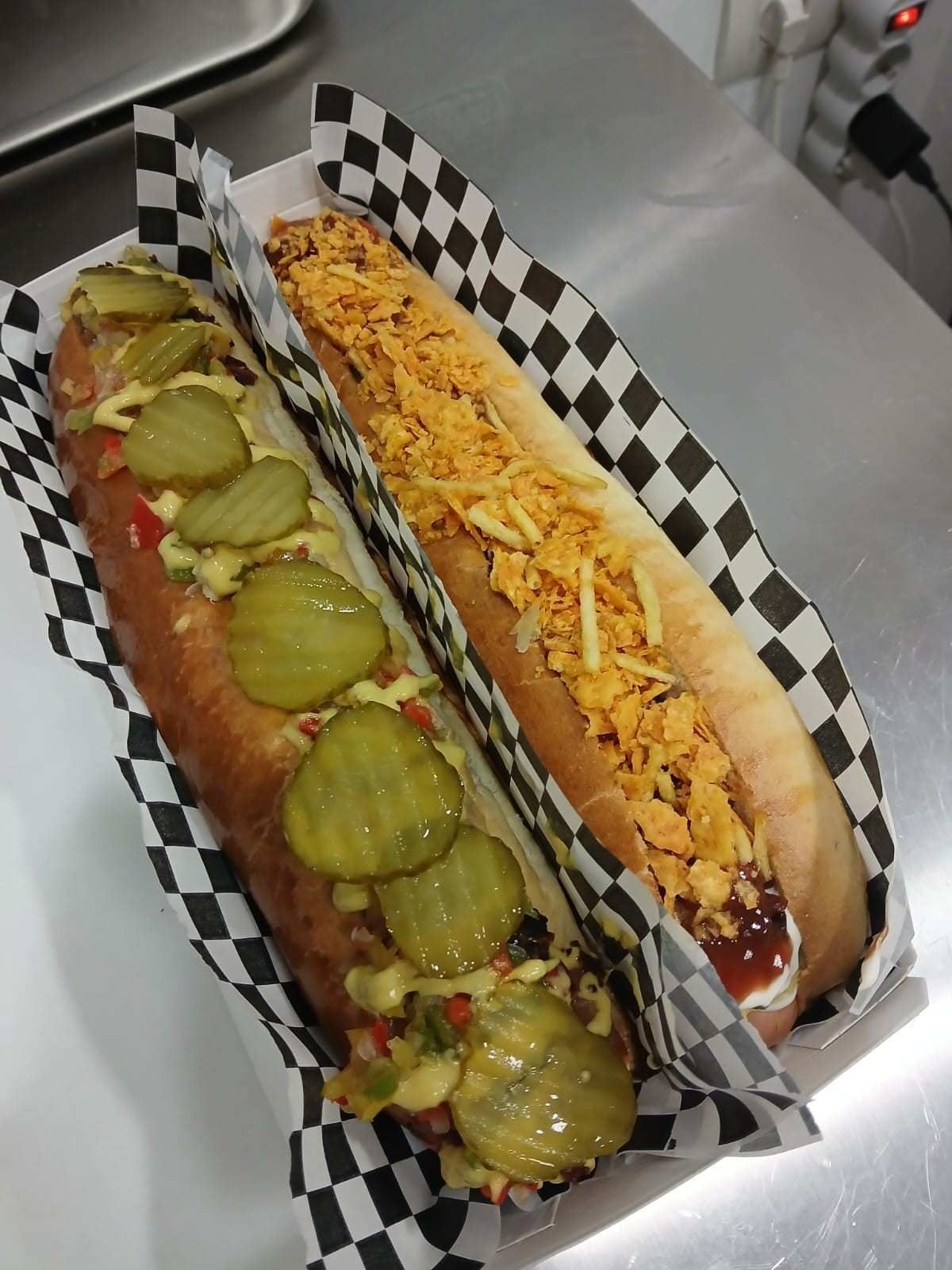 Dos perritos calientes grandes en envases de papel a cuadros. Uno con pepinillos y el otro con queso.