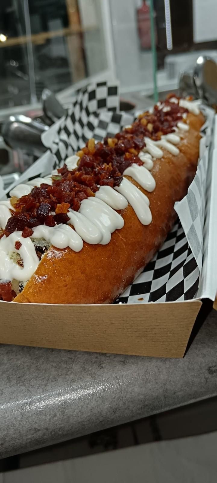 Primer plano de un hot dog largo y dorado con tocino, salsa blanca y un papel a cuadros blanco y negro.