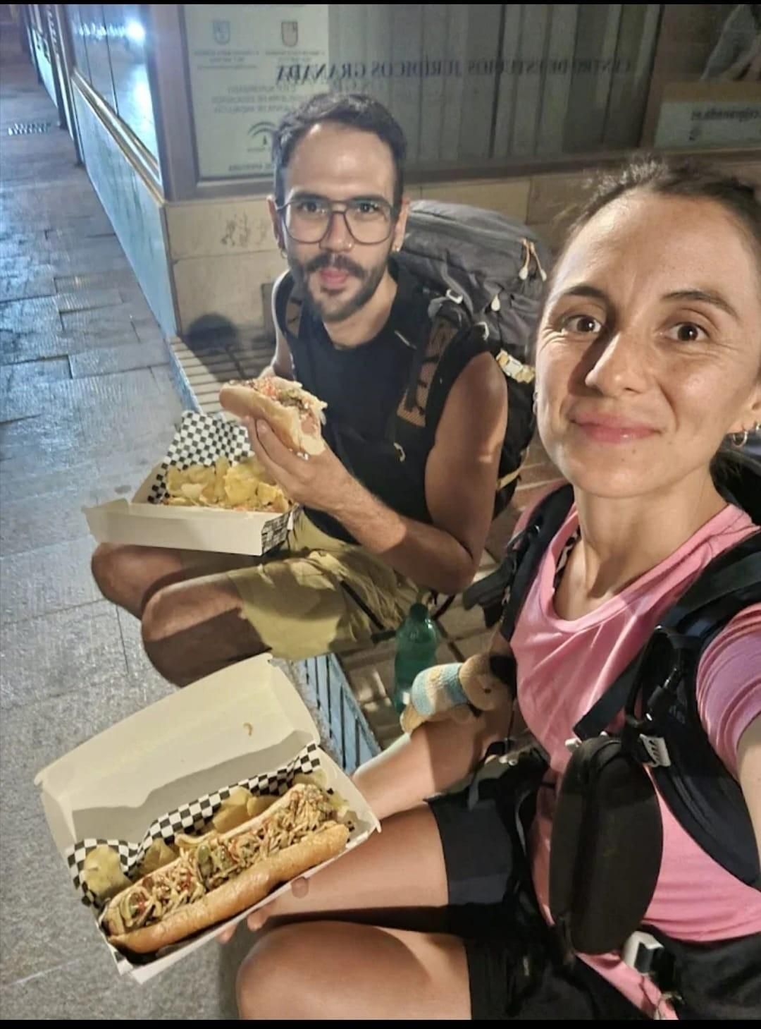 Dos personas vestidas con ropa deportiva comen perritos calientes y patatas fritas al aire libre.