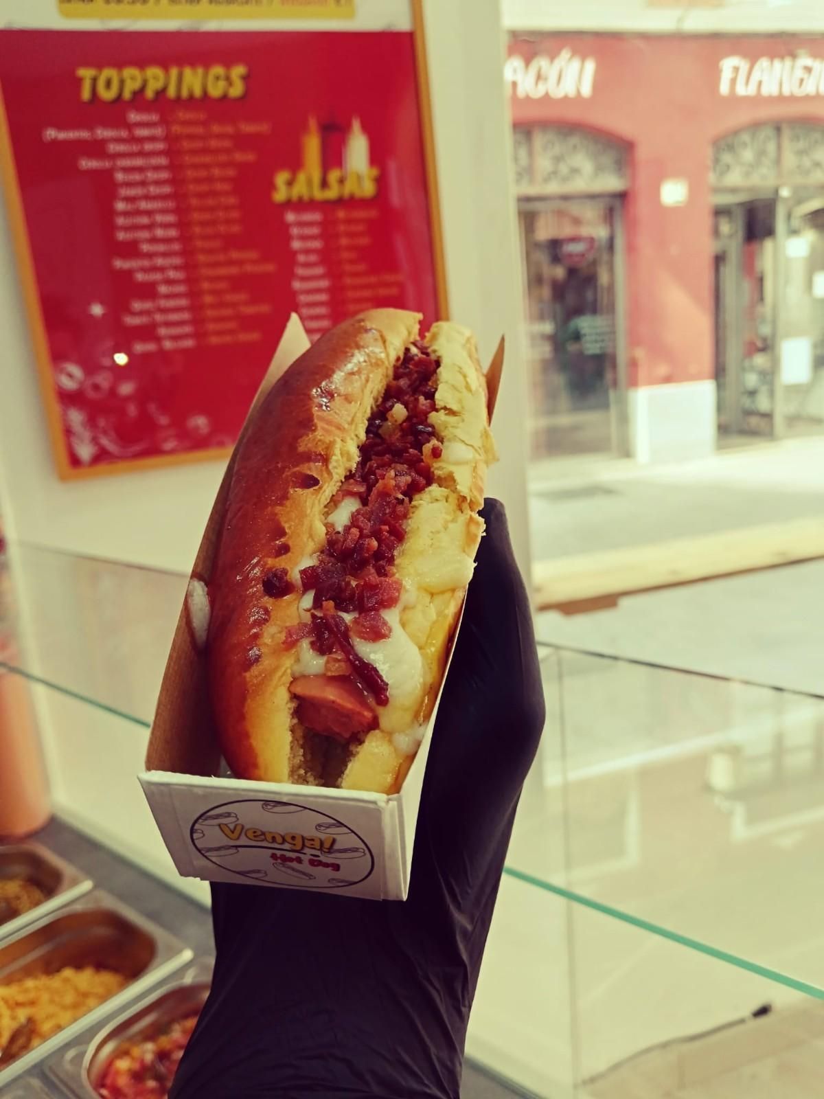 Perro caliente en una caja sostenido por una mano enguantada, con un menú de aderezos y salsas en el fondo.