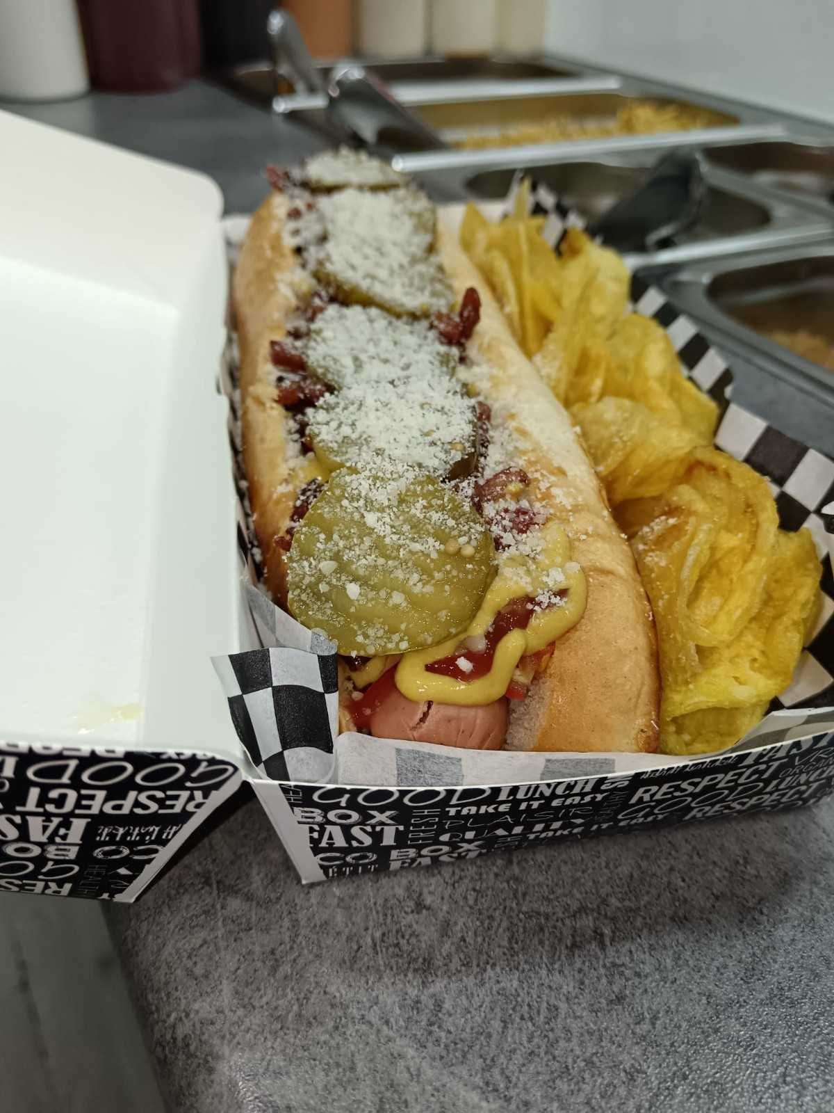 Perro caliente con pepinillos, mostaza y patatas fritas en una bandeja de papel.