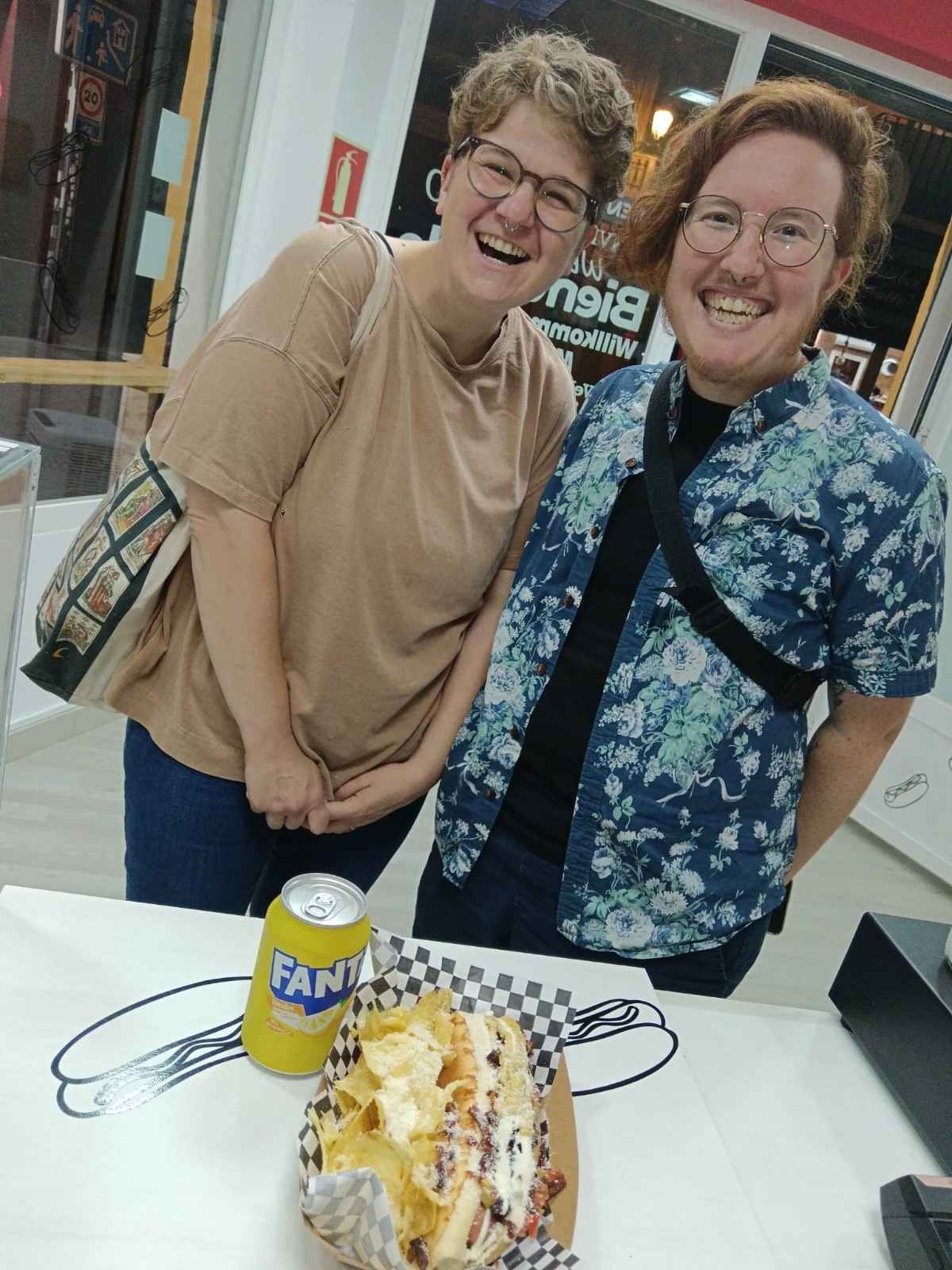 Dos personas se ríen tras una mesa con comida y un refresco. Una lleva gafas y una camisa floreada.