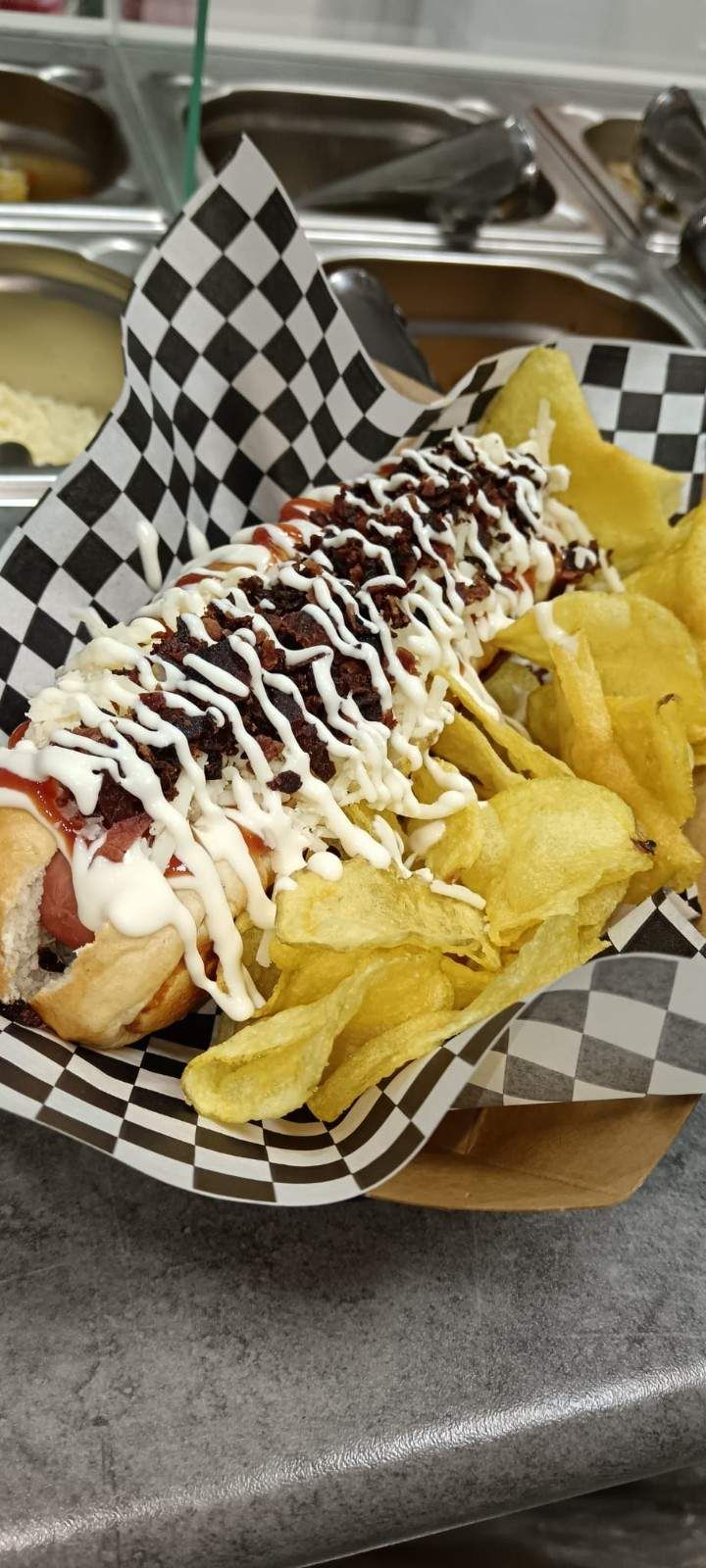 Un hot dog gourmet con patatas fritas en una cesta a cuadros, cubierto con salsa blanca y trocitos de tocino.