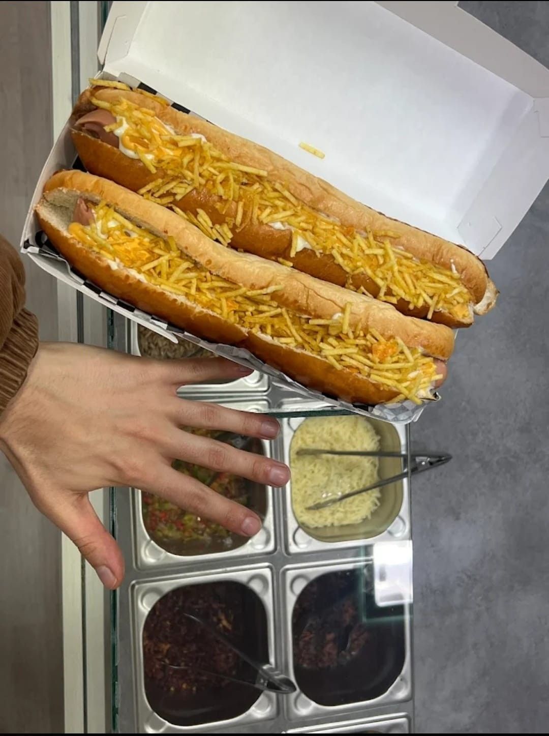 Dos perritos calientes largos cubiertos con patatas ralladas, en una caja abierta. Una mano señala cerca de los ingredientes.