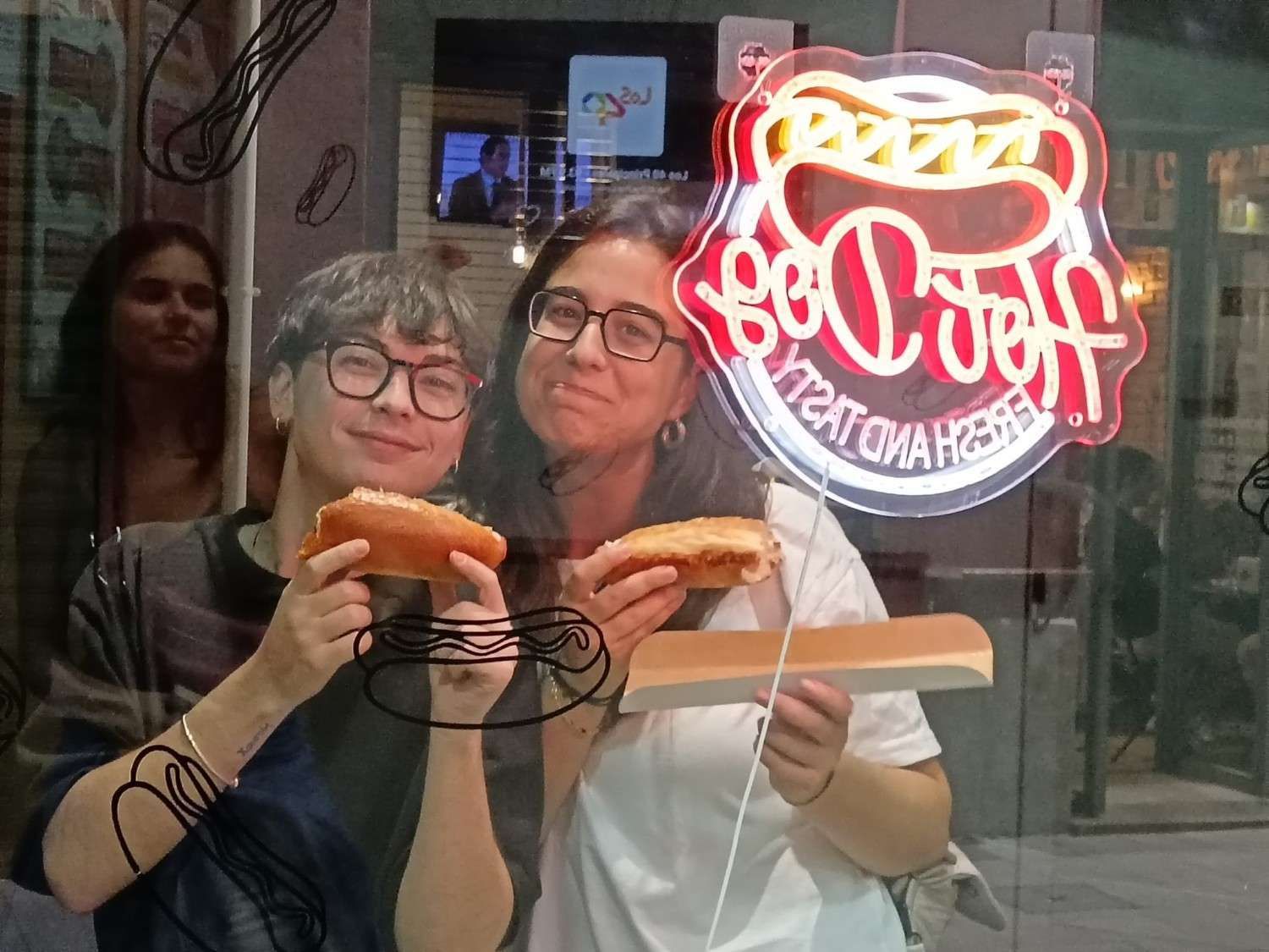 Dos personas sosteniendo perritos calientes, sonriendo frente a un cartel de neón que dice 