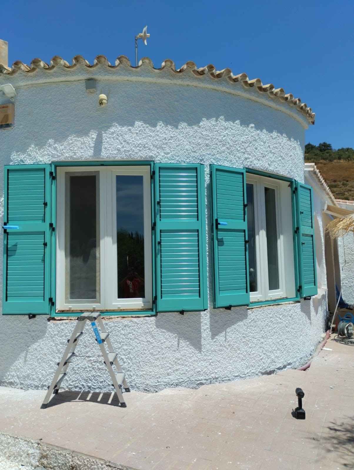 Pequeña casa blanca de estuco con contraventanas turquesas y techo curvo en un día soleado.