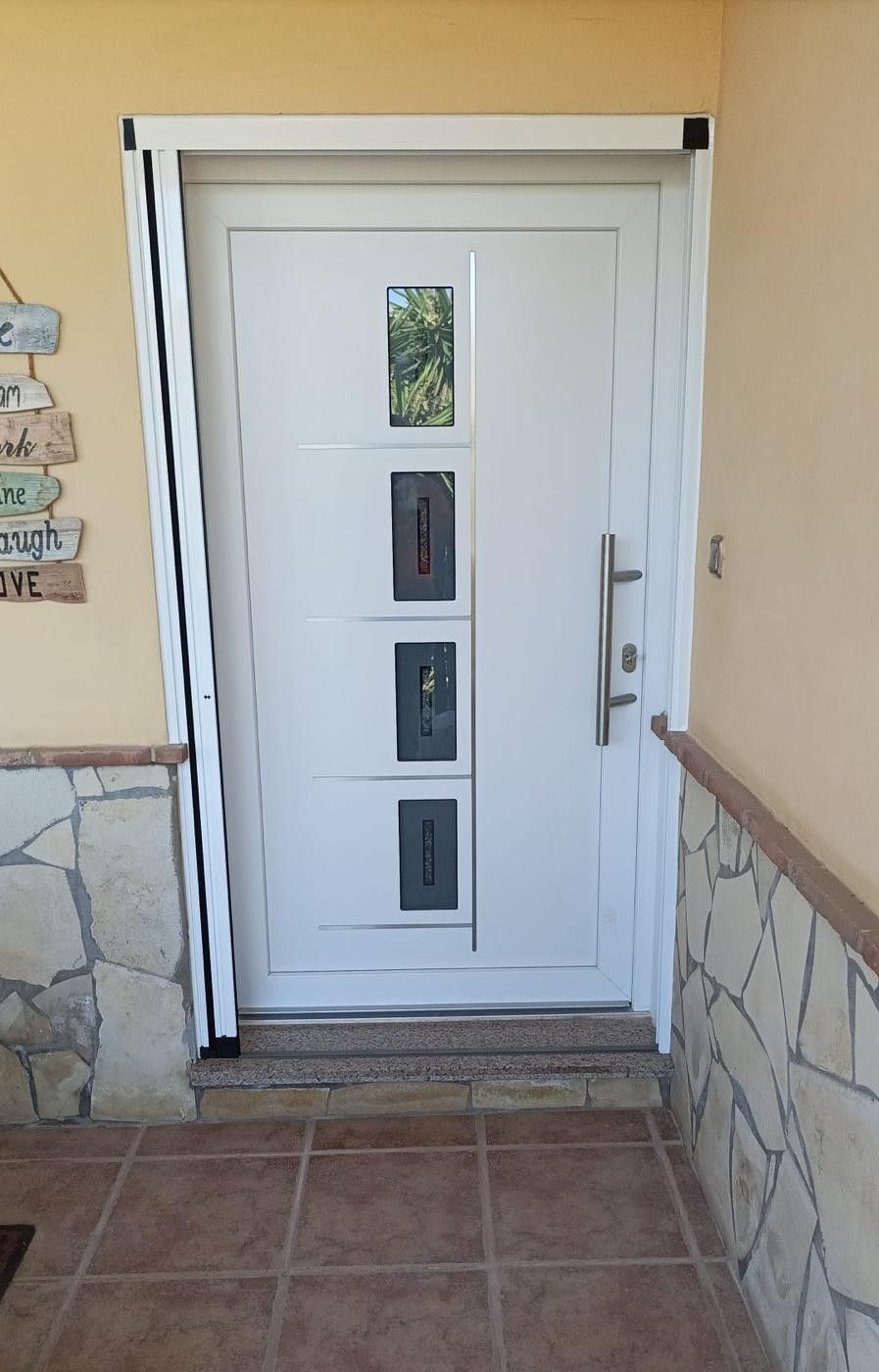 Puerta principal blanca con ventanas estrechas, manija plateada, revestimiento de piedra y entrada con azulejos.