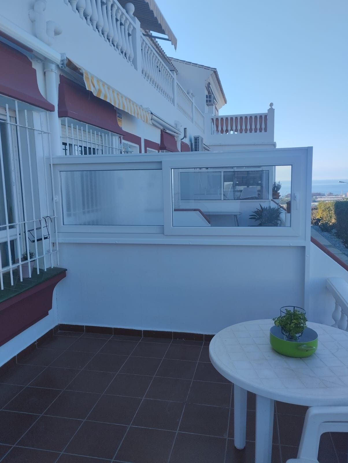 Balcón con barandilla blanca, suelo de baldosas, mesa redonda y vistas a la costa más allá del edificio.