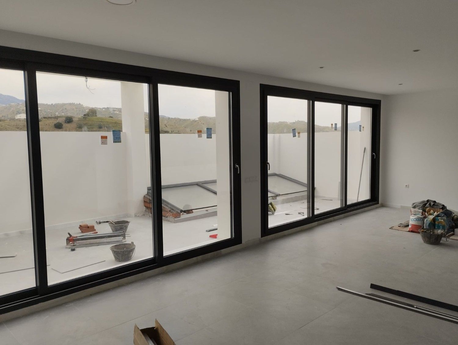 Habitación moderna y vacía con grandes ventanales con marcos negros y suelo sin terminar, con vistas a una obra en construcción.