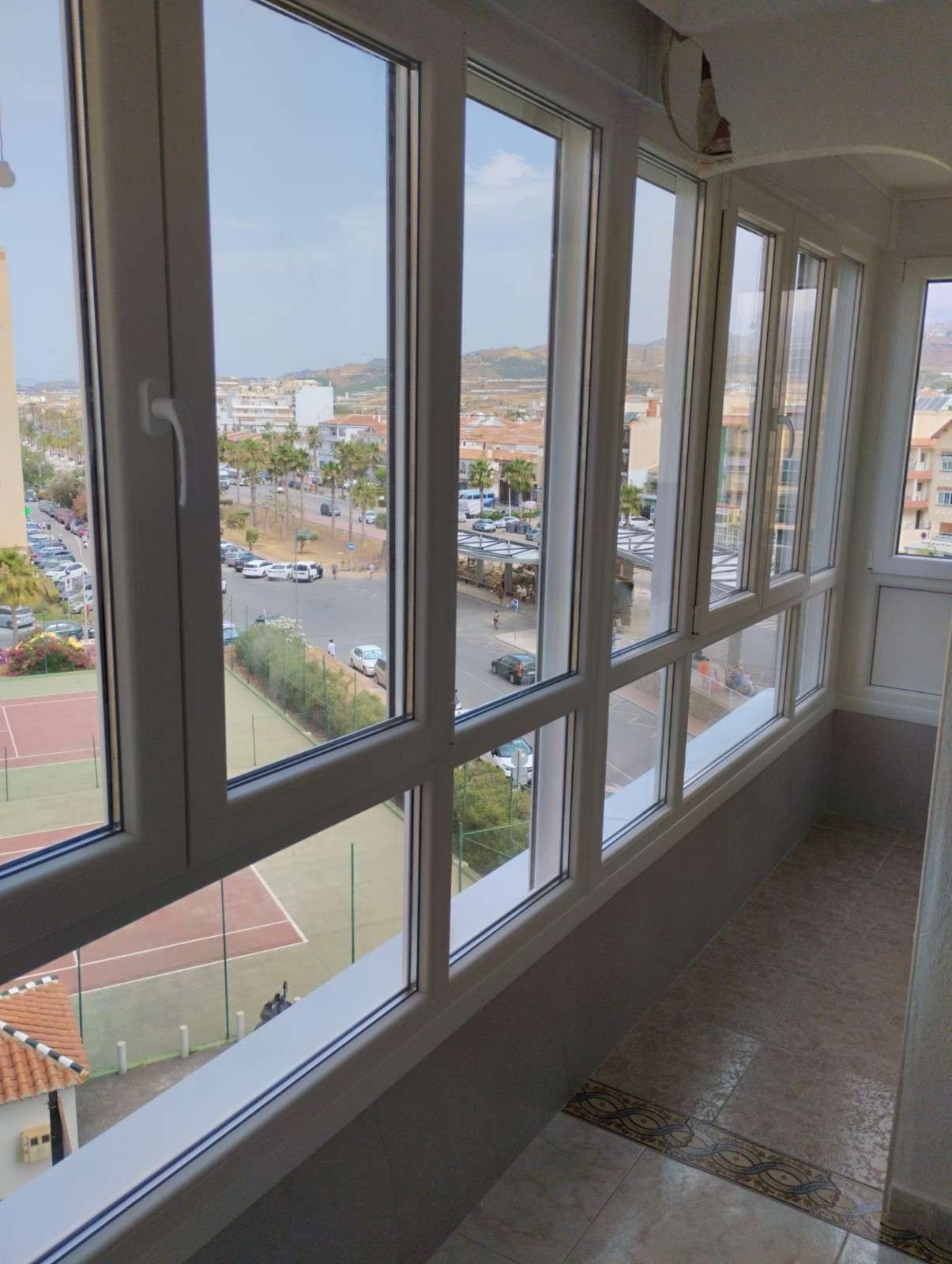 Pasillo luminoso con grandes ventanales con vistas a la ciudad y a una pista deportiva.