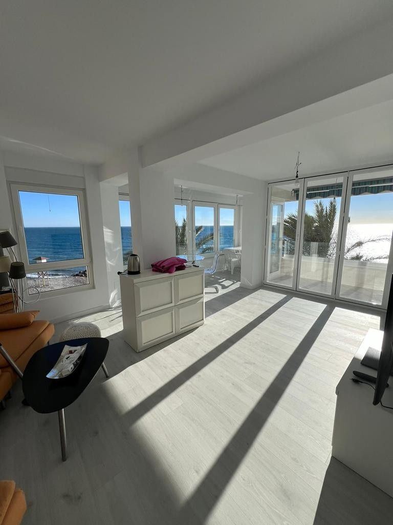 Habitación luminosa frente al mar con muebles blancos, puertas corredizas de vidrio y largas sombras proyectadas por el sol de la mañana.