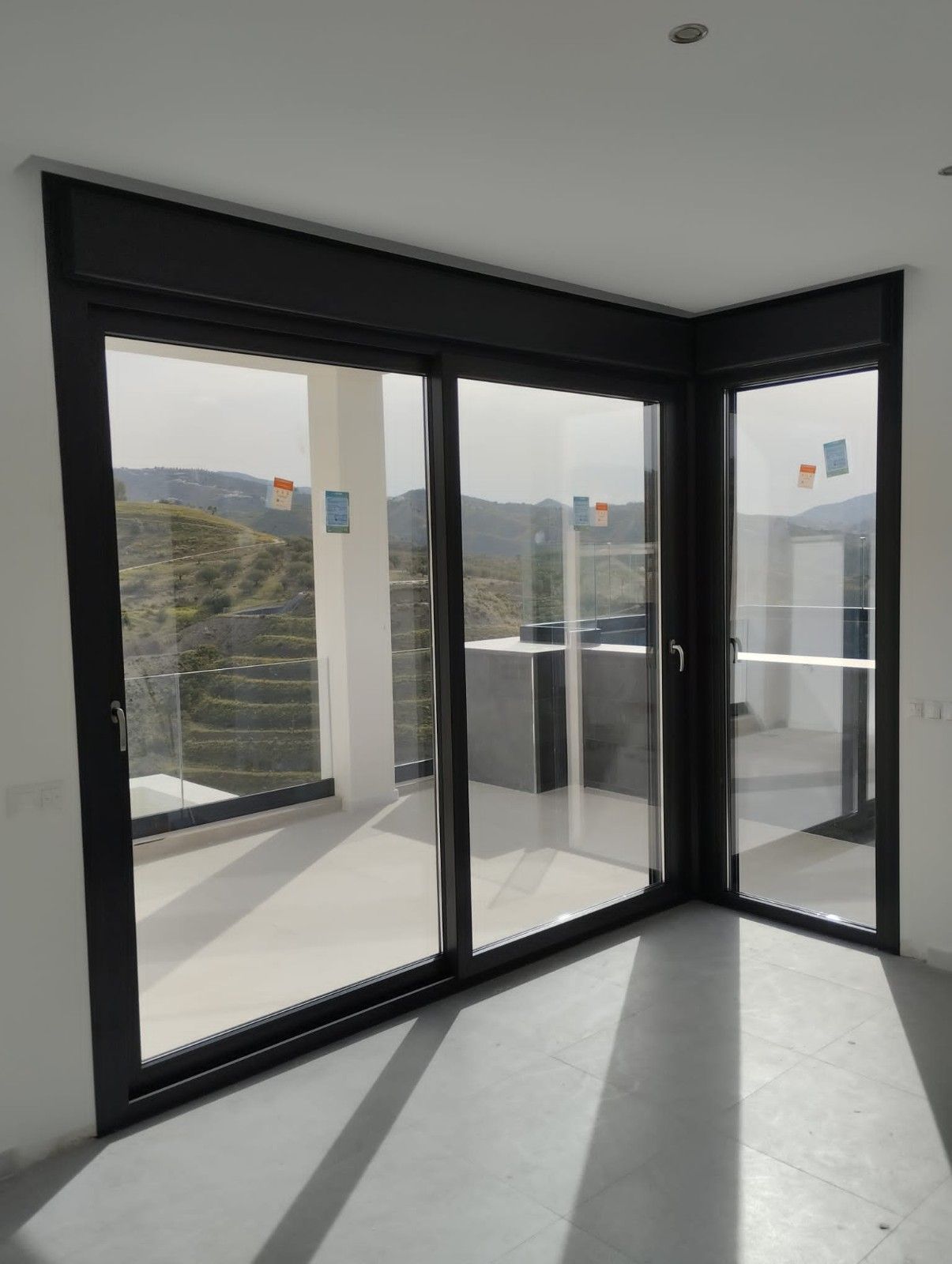 Habitación moderna y vacía con ventanas de esquina de cristal con marcos negros y suelo de hormigón bañado por el sol.