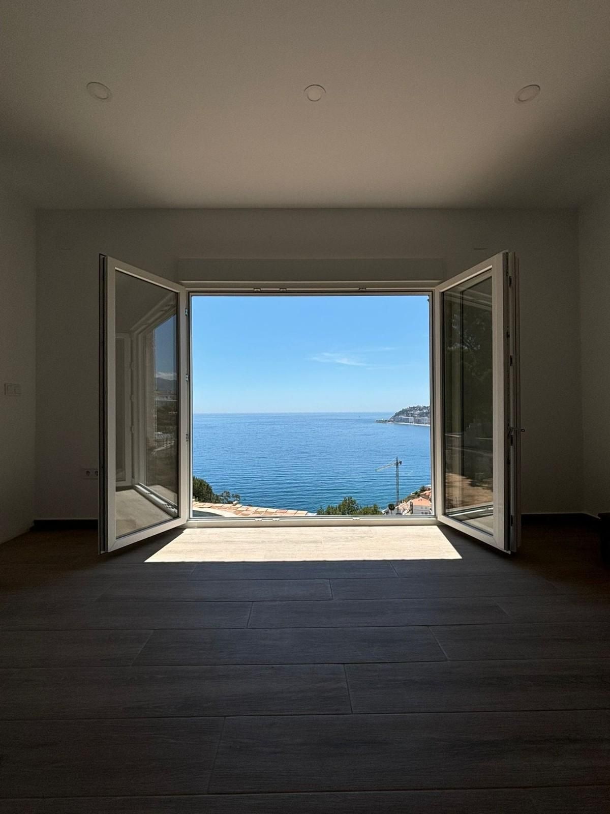 Puertas blancas abiertas que enmarcan una luminosa vista al océano desde una habitación interior oscura.