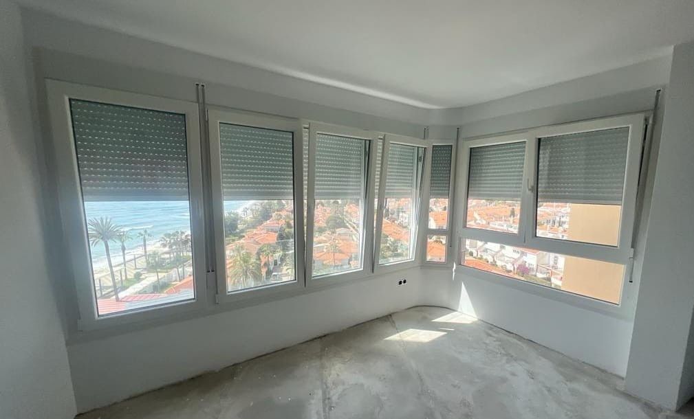 Habitación luminosa y vacía con grandes ventanales, paredes blancas y vistas a árboles y edificios en el exterior.